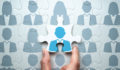 A puzzle made up of the grey silhouettes of businesspeople. A hand is placing the final puzzle piece, which has a blue silhouette