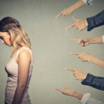 Is junior capital PIK’s innocent victim? Concept of accusation guilty person girl. Side profile sad upset woman looking down many fingers pointing at her back isolated on grey office wall background. Human face expression emotion feeling