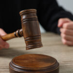 Judge with gavel sitting at wooden table against light grey background, closeup