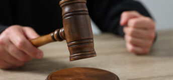 Judge with gavel sitting at wooden table against light grey background, closeup