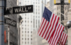 Wall street sign in New York City with American flags and New York Stock Exchange in background.